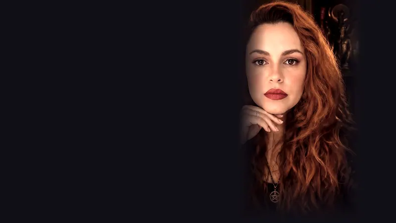 Retrato da Adséia à direita, cabelo ruivo longo, batom vermelho, mão no queixo, colar com pentagrama, ambiente escuro com velas